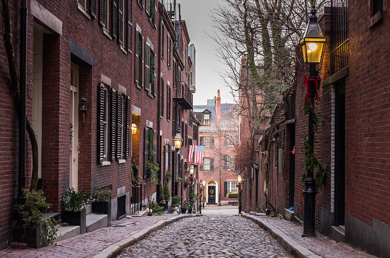 The narrow cobblestone lane of Acorn Street on Beacon Hill, lined with brick rowhouses and gas lamps.